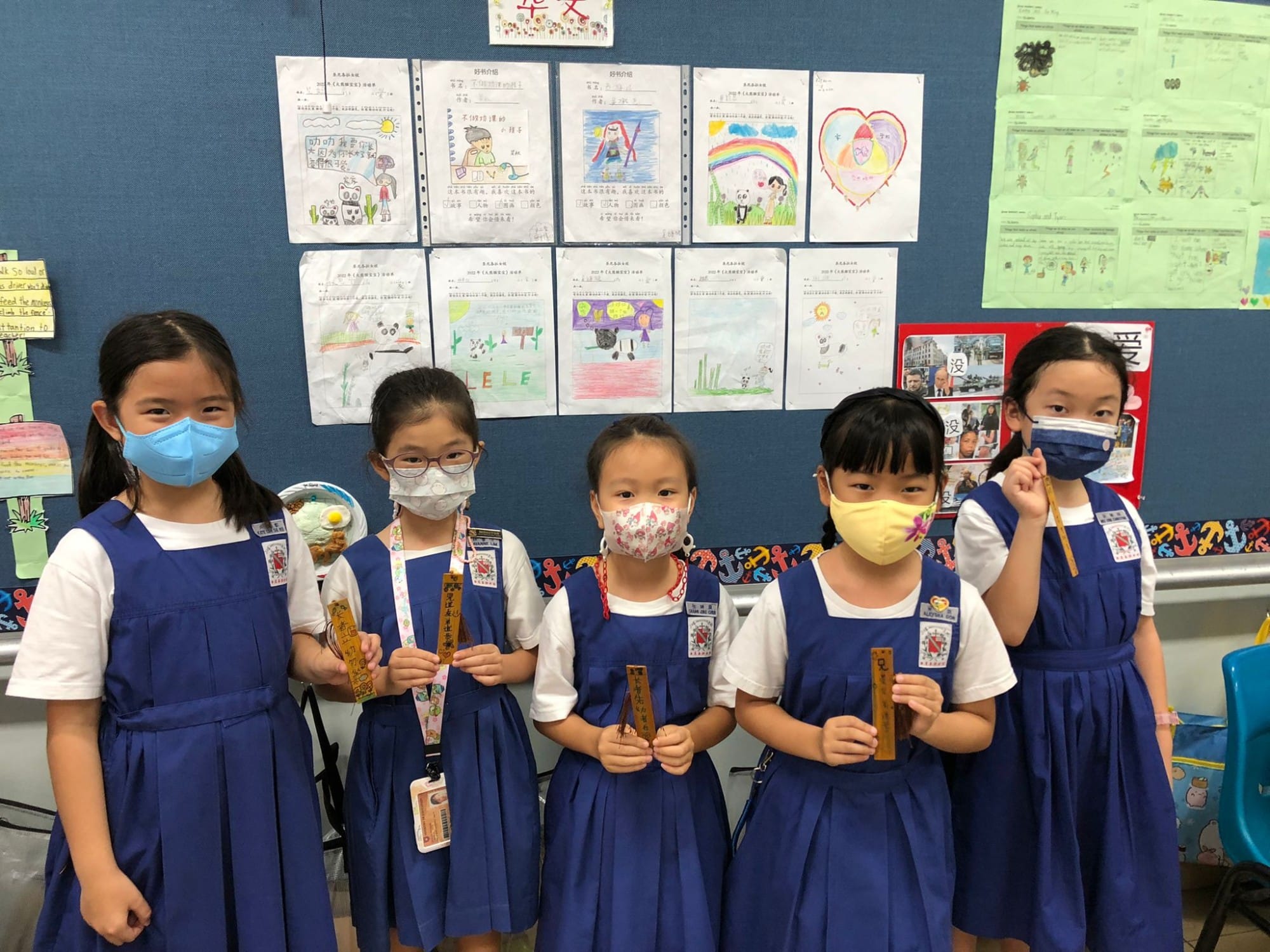 Five young girls in blue uniforms and face masks hold wooden bookmarks in front of a bulletin board with children's artwork.